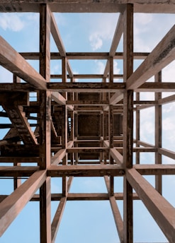 A complex structure of wooden beams forms a geometric grid pattern, viewed from below against a backdrop of a bright blue sky with scattered white clouds. The beams display a weathered texture, with an intricate interplay of light and shadow, creating a sense of depth and perspective.