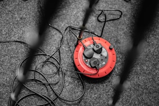 A red round electrical extension cord reel with multiple black plugs connected, resting on a dark gray carpeted floor. Several cables are sprawling outwards, some crossing each other.