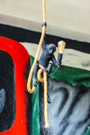 A black monkey figure is gripping a yellow rope while holding a light bulb in one hand. The background features a colorful mural with abstract shapes and colors like red, green, and black.