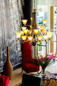 A sophisticated interior space featuring a large, elegant chandelier with numerous yellow lights arranged in a stylish pattern. Below the chandelier is a high-backed, plush red chair positioned next to a glass window. The surroundings include a textured, dark wall with a forest mural and a woven, vase-shaped decorative piece. In the foreground, a dark wooden table with a unique design supports a vase with greenery.