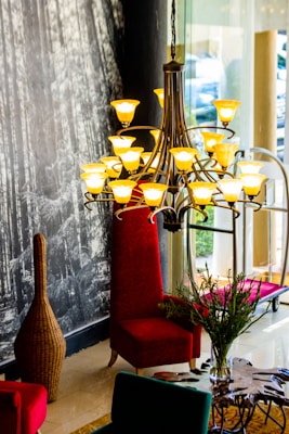 A sophisticated interior space featuring a large, elegant chandelier with numerous yellow lights arranged in a stylish pattern. Below the chandelier is a high-backed, plush red chair positioned next to a glass window. The surroundings include a textured, dark wall with a forest mural and a woven, vase-shaped decorative piece. In the foreground, a dark wooden table with a unique design supports a vase with greenery.