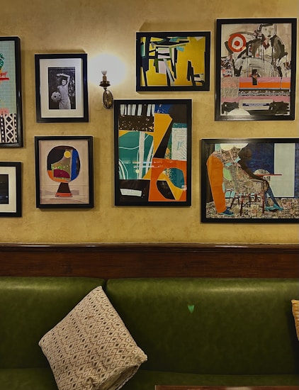 A collection of framed artworks arranged on a wall featuring abstract designs and a monochrome portrait. The wall is illuminated by a sconce above a green upholstered couch with a patterned cushion.
