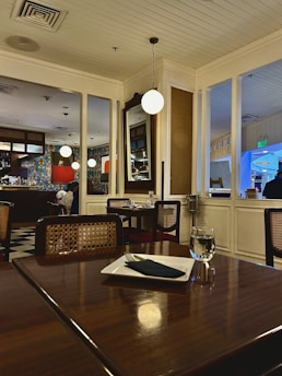 An elegant restaurant interior features a dark wooden table set with a white plate and black napkin, next to a glass of water. The chairs have cane backs, adding a classic touch. The restaurant has a ceiling with wooden panels and warm lighting hung from above. Mirrors and decoratively patterned walls can be seen, and a few individuals are seated in the background, possibly diners.