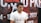 A person wearing a white t-shirt with the word 'TRAVE' partially visible is seated in front of a backdrop with text in large, bold letters and numbers. The backdrop includes information such as a prize pool mention and a date in 2023, featuring a predominantly red and black color scheme.