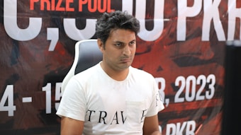 A person wearing a white t-shirt with the word 'TRAVE' partially visible is seated in front of a backdrop with text in large, bold letters and numbers. The backdrop includes information such as a prize pool mention and a date in 2023, featuring a predominantly red and black color scheme.