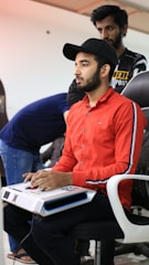A young man is sitting on a chair, focused intently on using a white arcade-style controller. He is wearing a bright red shirt and a black cap. Another person stands behind him observing. The setting appears to be a casual indoor environment, as indicated by people in the background and the relaxed atmosphere.