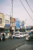A busy street scene featuring several people, motorbikes, and a van. The surroundings include multiple storefronts with colorful signage in an urban setting. There are various signboards and advertisements on the buildings, including those for BCA Syariah. A metal pole and wires can be seen overhead, and pedestrians are interacting with the vendors and street traffic.