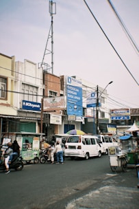 A busy street scene featuring several people, motorbikes, and a van. The surroundings include multiple storefronts with colorful signage in an urban setting. There are various signboards and advertisements on the buildings, including those for BCA Syariah. A metal pole and wires can be seen overhead, and pedestrians are interacting with the vendors and street traffic.