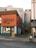 A small, colorful building with a red and blue exterior, likely a restaurant or cafe, featuring a sign with stylized lettering. Outside, there are a few high tables and stools, suggesting outdoor seating. The building is situated in an urban environment, with adjacent structures and narrow alleyways visible.