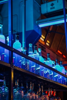 A collection of illuminated bottles is arranged on a shelf, with a neon blue light enhancing their translucent appearance. Below the bottles, rows of hanging wine glasses create a reflective and frosted look. The background features industrial elements and warm lighting, adding an urban atmosphere.