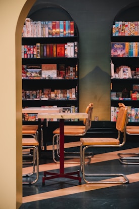 A cozy room with a wooden table surrounded by four chairs with wicker seats and backs, positioned at the center. Behind the table, a well-organized shelf filled with colorful board games suggests a relaxed and inviting atmosphere. The space is enclosed by an archway, adding an element of architectural interest.