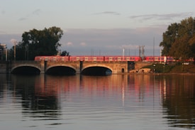 A red train travels across a multi-arched bridge over a calm river, reflecting the train and evening sky on the water's surface. The bridge is surrounded by lush green trees and a few people can be seen near the shoreline enjoying the serene setting.