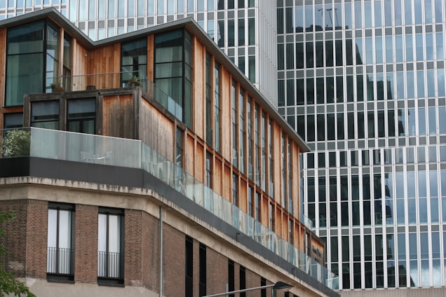 A modern architectural building with a combination of wood and glass materials. The facade features large windows and a glass balcony, creating a sleek and contemporary look. The background displays the reflective glass surface of a taller building.