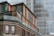 A modern architectural building with a combination of wood and glass materials. The facade features large windows and a glass balcony, creating a sleek and contemporary look. The background displays the reflective glass surface of a taller building.