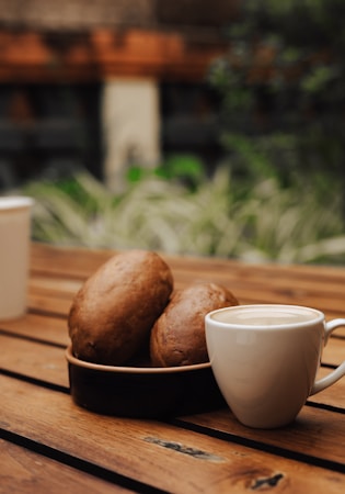 A cozy setting featuring a wooden table with a white cup filled with coffee alongside a plate holding two round, brown bread rolls. The background shows blurred green foliage and a stone or brick structure, adding to the warm, inviting atmosphere.