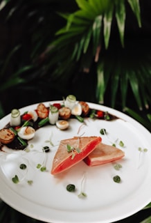 A beautifully plated dish featuring two pieces of seared fish with a pink center, garnished with microgreens. Surrounding the fish are small scallops, slices of cucumber, green beans, quail eggs, and red cherry tomatoes, all arranged artistically on a white ceramic plate. The background consists of lush green foliage.