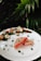 A beautifully plated dish featuring two pieces of seared fish with a pink center, garnished with microgreens. Surrounding the fish are small scallops, slices of cucumber, green beans, quail eggs, and red cherry tomatoes, all arranged artistically on a white ceramic plate. The background consists of lush green foliage.