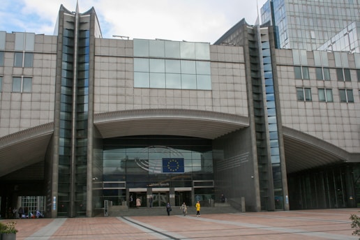 A large, modern building with a prominent entrance displaying the European Union flag. The architecture features tall columns and a symmetrical design. Several people can be seen walking near the entrance.
