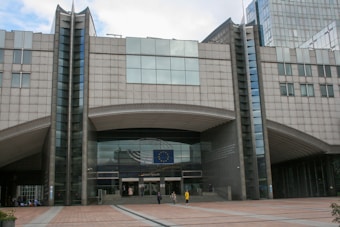 A large, modern building with a prominent entrance displaying the European Union flag. The architecture features tall columns and a symmetrical design. Several people can be seen walking near the entrance.