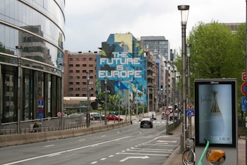 An urban street scene with a large building mural reading 'The Future is Europe.' Modern architecture surrounds the area with a mix of office buildings and residential structures. There are several street signs, an advertisement for a fragrance, and a bike parked on the sidewalk. The street is mostly clear with a few cars and pedestrians visible.
