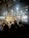 A live music event with a band performing on stage. Bright lights and multimedia screens illuminate the space, creating a lively atmosphere. The audience is standing with several people raising their hands, suggesting engagement and enthusiasm.
