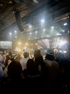 A live music event with a band performing on stage. Bright lights and multimedia screens illuminate the space, creating a lively atmosphere. The audience is standing with several people raising their hands, suggesting engagement and enthusiasm.
