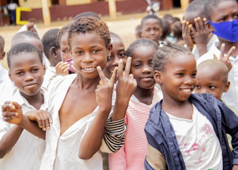 A group of smiling children are gathered closely together, some making peace signs with their hands. They are outdoors, possibly in a school setting, with a building visible in the background.