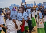 A group of children stands outside holding notebooks with covers showing a logo and photographs. They are wearing school uniforms consisting of white shirts and green shorts or skirts. The setting appears to be a schoolyard with buildings and a clear sky in the background.