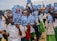 A group of children stands outside holding notebooks with covers showing a logo and photographs. They are wearing school uniforms consisting of white shirts and green shorts or skirts. The setting appears to be a schoolyard with buildings and a clear sky in the background.