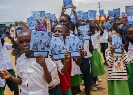 A group of children stands outside holding notebooks with covers showing a logo and photographs. They are wearing school uniforms consisting of white shirts and green shorts or skirts. The setting appears to be a schoolyard with buildings and a clear sky in the background.