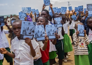 A group of children stands outside holding notebooks with covers showing a logo and photographs. They are wearing school uniforms consisting of white shirts and green shorts or skirts. The setting appears to be a schoolyard with buildings and a clear sky in the background.
