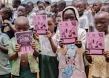 A group of children holding pink notebooks in a crowded setting. The children are wearing various school uniforms and casual clothing. The background shows more children and part of a building structure.