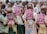 A group of children holding pink notebooks in a crowded setting. The children are wearing various school uniforms and casual clothing. The background shows more children and part of a building structure.
