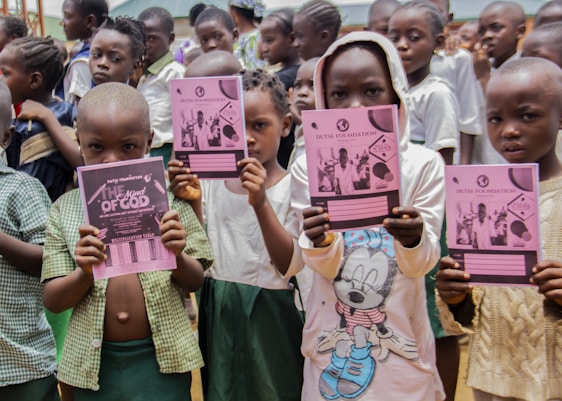 A group of children holding pink notebooks in a crowded setting. The children are wearing various school uniforms and casual clothing. The background shows more children and part of a building structure.