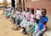 A group of children sitting in a row on a concrete step, each holding a booklet. The children are wearing a school uniform of green checked dresses, with the exception of one child wearing a blue and orange t-shirt. They are seated outside a school building with a brown and yellow exterior.