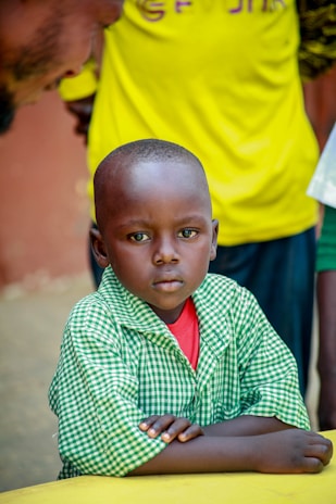 A young child with a serious expression, wearing a green checkered shirt over a red undershirt, is sitting with folded arms on a yellow surface. An adult is partially visible behind, wearing a bright yellow shirt.