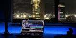 A laptop displaying coding software rests on a desk illuminated by a soft blue light. In the background, a cityscape is visible through a large window, with buildings lit up against the night sky. The scene is calm and focused, emphasizing a workspace oriented towards programming.