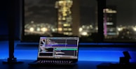 A laptop displaying coding software rests on a desk illuminated by a soft blue light. In the background, a cityscape is visible through a large window, with buildings lit up against the night sky. The scene is calm and focused, emphasizing a workspace oriented towards programming.