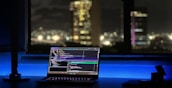 A laptop displaying coding software rests on a desk illuminated by a soft blue light. In the background, a cityscape is visible through a large window, with buildings lit up against the night sky. The scene is calm and focused, emphasizing a workspace oriented towards programming.
