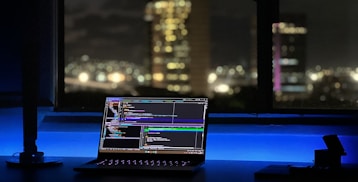 A laptop displaying coding software rests on a desk illuminated by a soft blue light. In the background, a cityscape is visible through a large window, with buildings lit up against the night sky. The scene is calm and focused, emphasizing a workspace oriented towards programming.