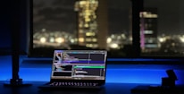 A laptop displaying coding software rests on a desk illuminated by a soft blue light. In the background, a cityscape is visible through a large window, with buildings lit up against the night sky. The scene is calm and focused, emphasizing a workspace oriented towards programming.