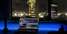 A laptop displaying coding software rests on a desk illuminated by a soft blue light. In the background, a cityscape is visible through a large window, with buildings lit up against the night sky. The scene is calm and focused, emphasizing a workspace oriented towards programming.