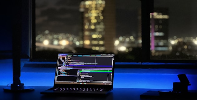 A laptop displaying coding software rests on a desk illuminated by a soft blue light. In the background, a cityscape is visible through a large window, with buildings lit up against the night sky. The scene is calm and focused, emphasizing a workspace oriented towards programming.
