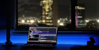 A laptop displaying coding software rests on a desk illuminated by a soft blue light. In the background, a cityscape is visible through a large window, with buildings lit up against the night sky. The scene is calm and focused, emphasizing a workspace oriented towards programming.