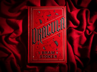 A bright red book cover with ornate black gothic lettering that reads 'Dracula' by Bram Stoker. The book is placed on a textured red fabric, creating a dramatic and moody aesthetic. The cover features decorative elements such as swirls and bat motifs in a dark color.