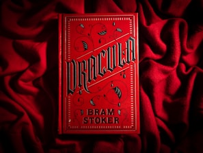 A bright red book cover with ornate black gothic lettering that reads 'Dracula' by Bram Stoker. The book is placed on a textured red fabric, creating a dramatic and moody aesthetic. The cover features decorative elements such as swirls and bat motifs in a dark color.