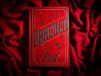 A bright red book cover with ornate black gothic lettering that reads 'Dracula' by Bram Stoker. The book is placed on a textured red fabric, creating a dramatic and moody aesthetic. The cover features decorative elements such as swirls and bat motifs in a dark color.