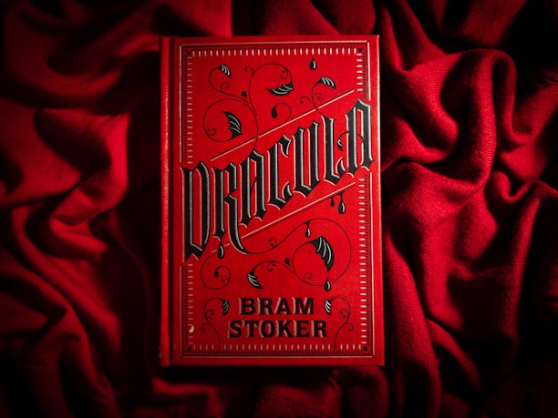 A bright red book cover with ornate black gothic lettering that reads 'Dracula' by Bram Stoker. The book is placed on a textured red fabric, creating a dramatic and moody aesthetic. The cover features decorative elements such as swirls and bat motifs in a dark color.