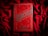 A bright red book cover with ornate black gothic lettering that reads 'Dracula' by Bram Stoker. The book is placed on a textured red fabric, creating a dramatic and moody aesthetic. The cover features decorative elements such as swirls and bat motifs in a dark color.
