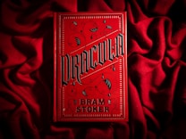 A bright red book cover with ornate black gothic lettering that reads 'Dracula' by Bram Stoker. The book is placed on a textured red fabric, creating a dramatic and moody aesthetic. The cover features decorative elements such as swirls and bat motifs in a dark color.